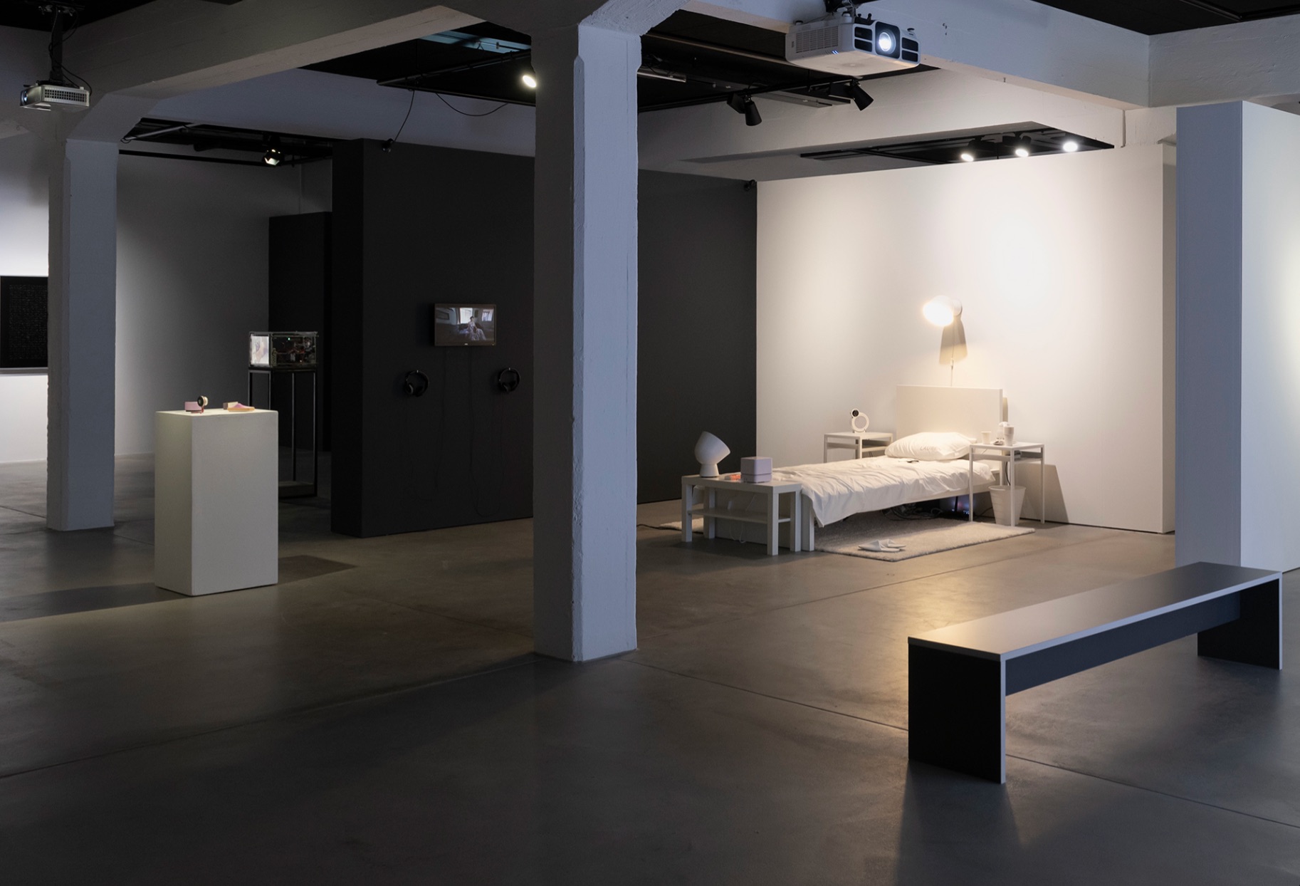 Gallery installation in a concrete-floored space: a single bed made up in white linen sits in a lit alcove with side tables, a lamp, a small camera and other domestic objects, while a wall-mounted screen and two pairs of headphones face it from a dark partition opposite.