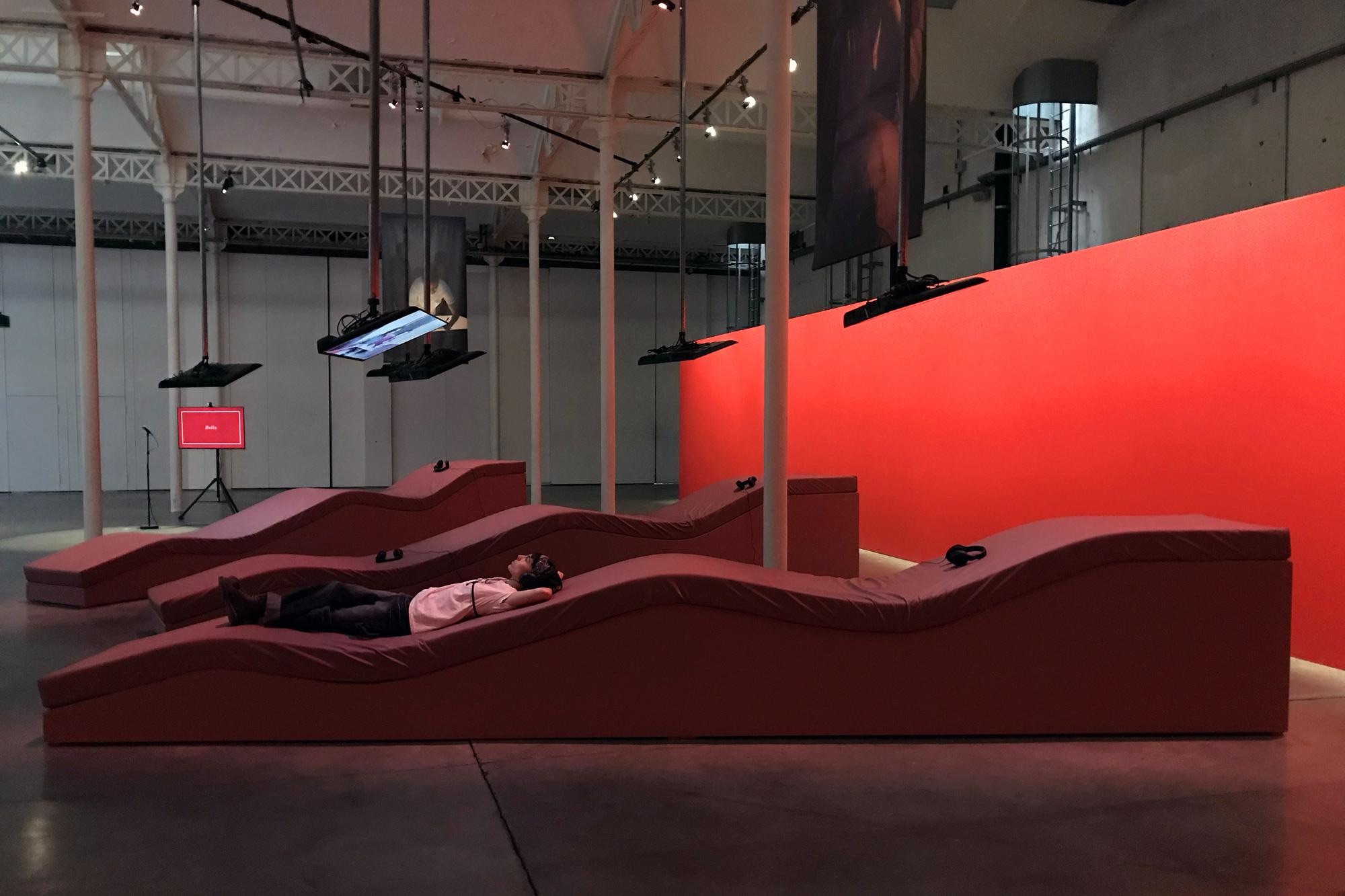 A person lies on their back wearing headphones on one of several large red wave-shaped foam platforms in an industrial hall, under suspended screens and beside a tall red-lit wall.