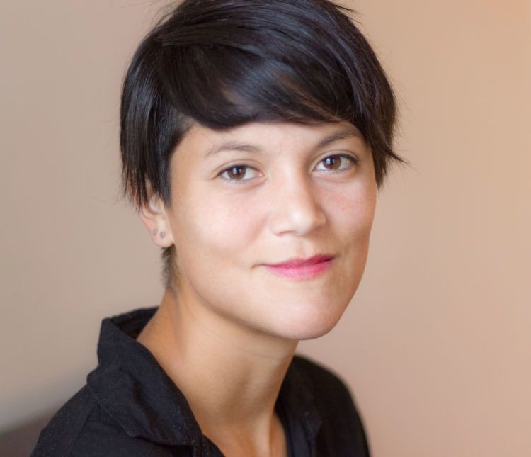 Head-and-shoulders portrait of Lauren Lee McCarthy, with short dark hair and a black shirt, looking towards the camera against a plain warm grey background.