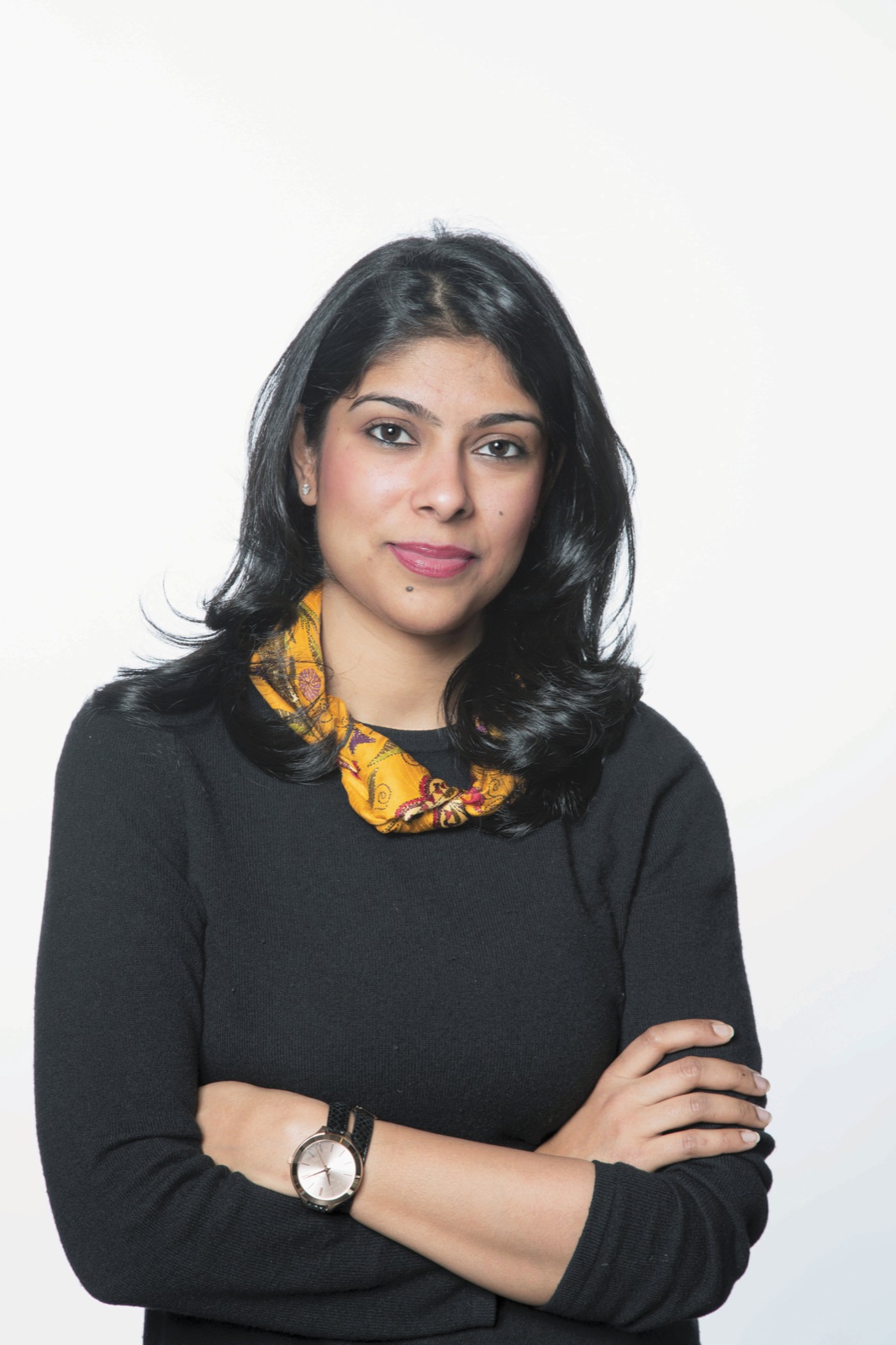 Manjari Sihare-Sutin standing with arms crossed, wearing a black sweater and an orange patterned scarf, against a plain white background.