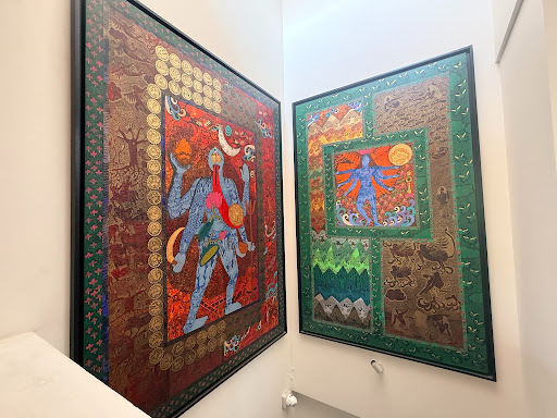 Two large framed tapestry-style works hung on adjoining gallery walls: a blue many-armed figure on red, and a blue many-armed figure in a shrine-like niche on green, both bordered in gold and red patterning.