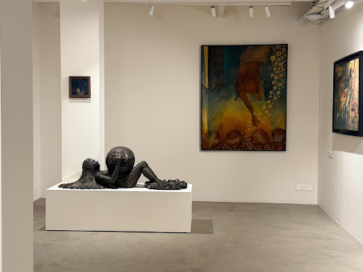 A gallery installation view: a dark bronze sculpture of a reclining figure with a spherical head on a low white plinth, with a large teal and gold painting on the wall behind it.