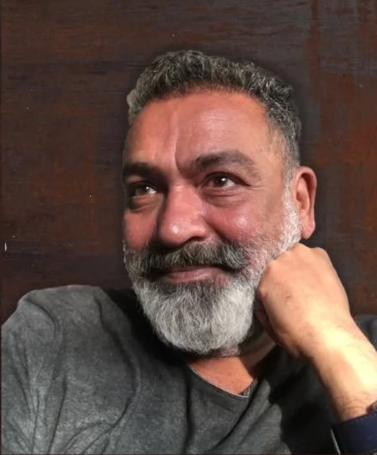 Somu Desai, chin resting on his hand, against a dark wall.