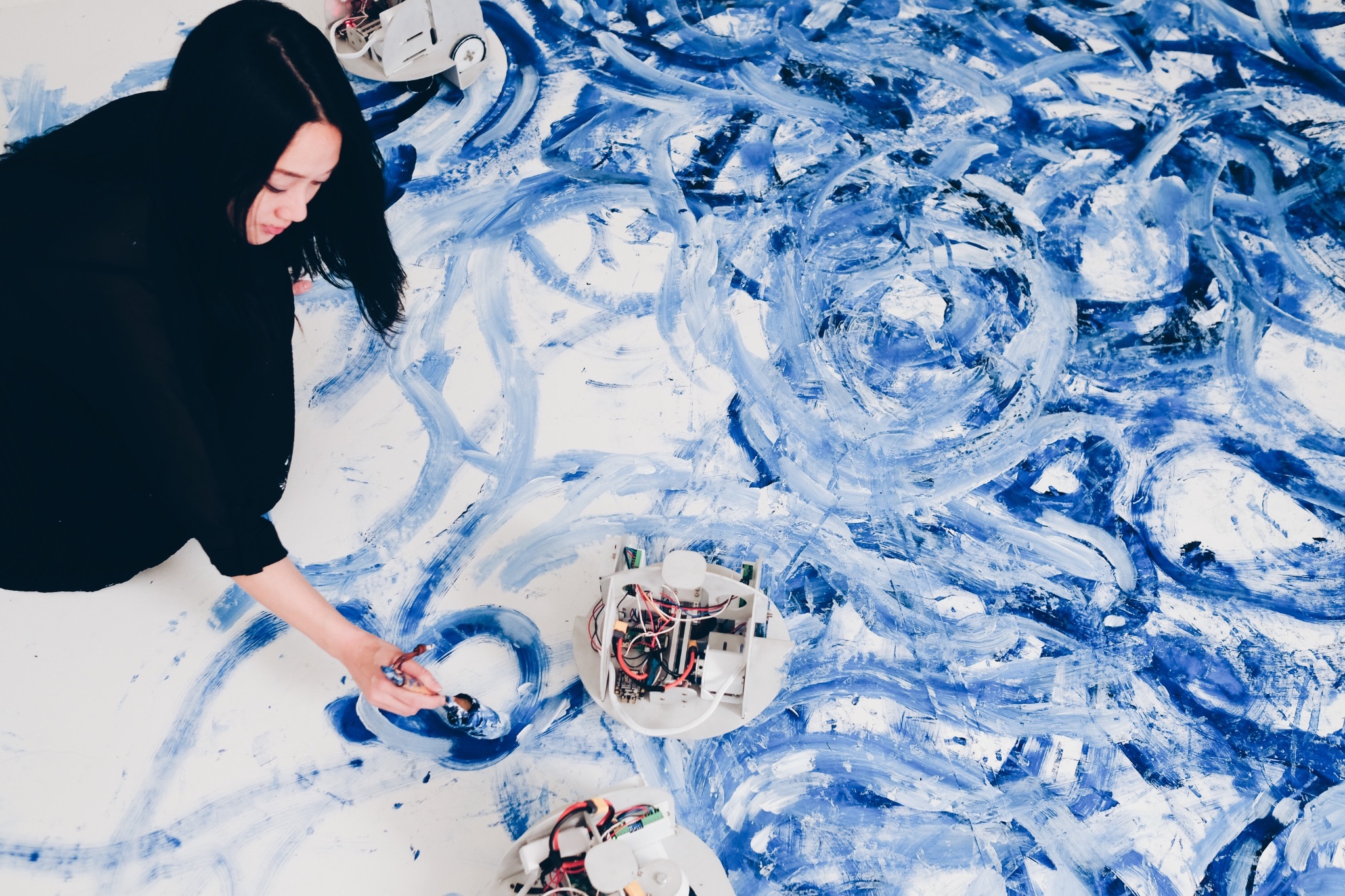 Overhead view of Sougwen Chung kneeling on a white floor painted with sweeping blue brushstrokes, drawing with a brush while three small wheeled robots carrying exposed circuit boards move across the same surface.