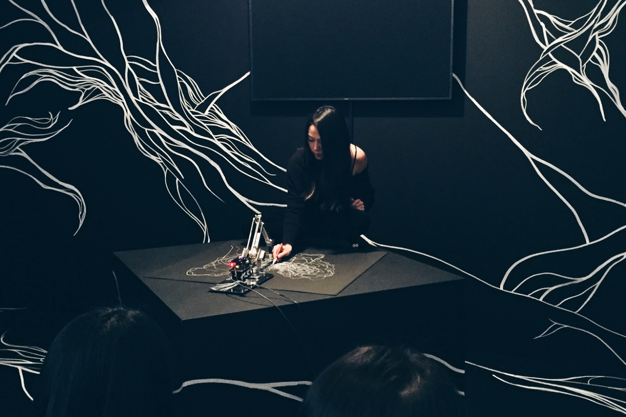 Chung seated at a low black plinth in a darkened room, drawing fine white lines beside a small robotic arm, with large white line drawings covering the surrounding black walls and the backs of two audience members in the foreground.